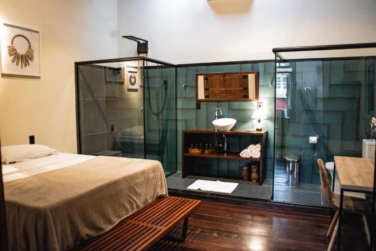 Una habitación de hotel moderna con cabina de ducha de vidrio, un banco de madera, una cama, una mesita y un lavamanos con espejo.
