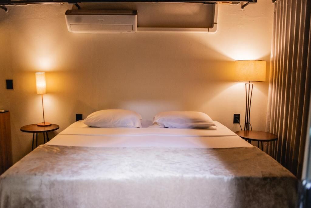 Een hotelkamer met een bed, twee bedlampen, een wandairconditioner en een gordijn.