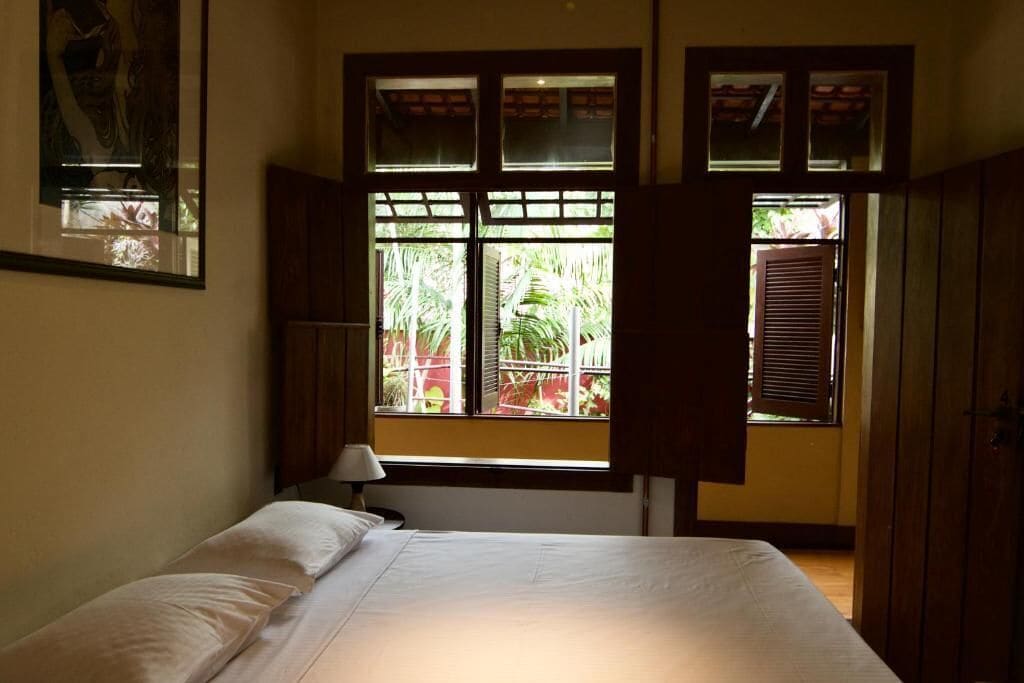 A bedroom with a bed, a window with shutters, a nightstand, and a wall-mounted lamp.