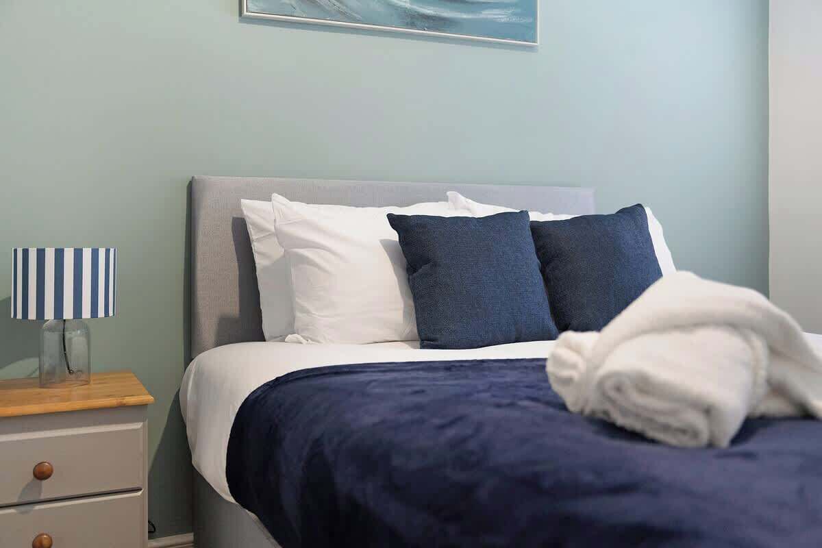 Een net opgemaakt bed met wit en marineblauwe beddengoed, een houten nachtkastje met een gestreepte lamp en een lijst met een afbeelding aan de muur.