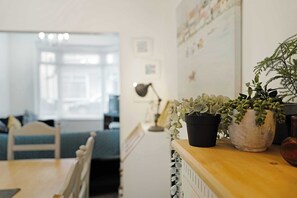 Una habitación con una mesa de madera, dos plantas en macetas, una lámpara y, al fondo, un sofá y un sillón azules.