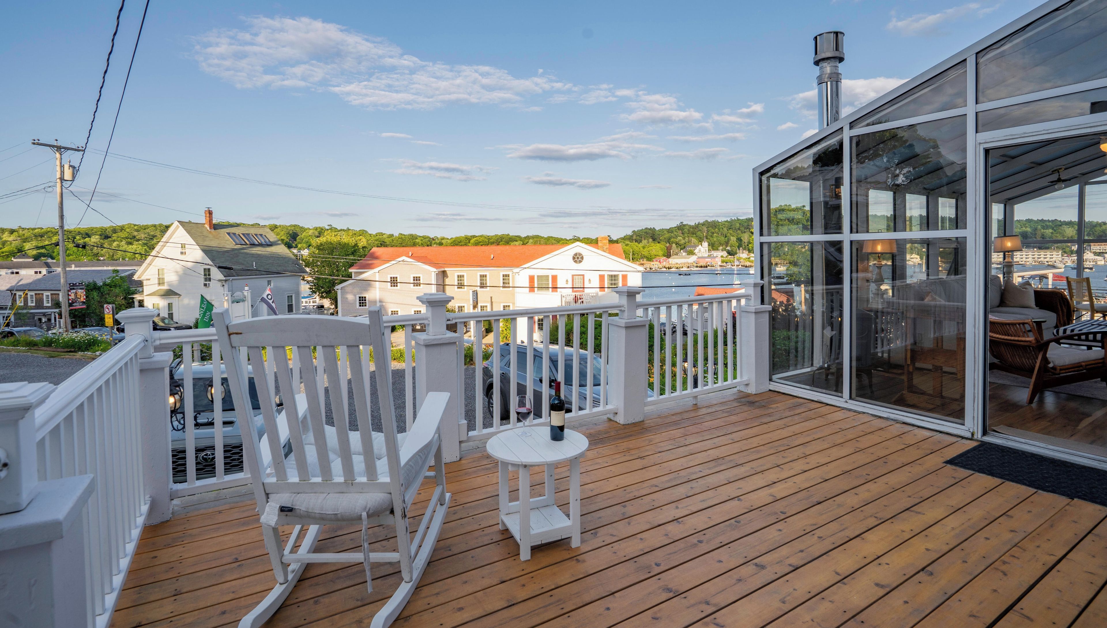 A wooden deck with a rocking chair and a small table, overlooking a town with buildings and a body of water.