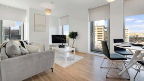 A modern living room with a sofa, TV, dining table, and chairs, large windows with a city view. - Manchester 2BR Apartment w/Super Fast WI-FI ! (Greater Manchester)