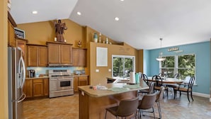 A spacious kitchen with wooden cabinets, a central island with bar stools, and a dining area with a blue wall and a 'gather' sign. - Eagle Ridge Retreat - Dock, Hot Tub & Game Room (Gravois Mills)