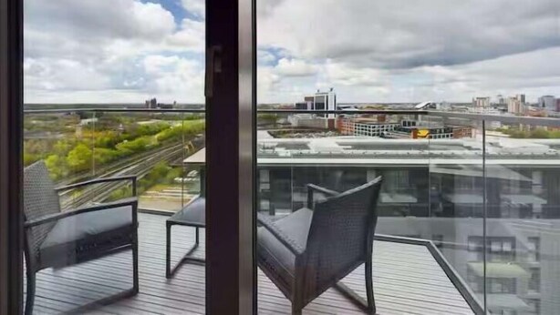 A balcony with two chairs and a glass railing overlooking a cityscape with buildings and a railway track. - 2 Bed Flat Near City Centre with Free Parking (Stretford)