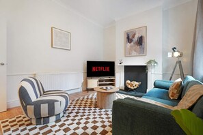 A living room with a striped armchair, a blue sofa, a coffee table, a fireplace, a TV displaying Netflix, and a framed abstract painting.