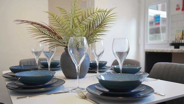 A dining table set with blue plates, bowls, and wine glasses, accompanied by a potted fern. - Charming 4BR Retreat in Hull | Sleeps 8 | by Stay With Us (Kingston upon Hull)