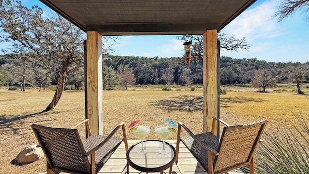 A covered outdoor seating area with two wicker chairs and a round table, overlooking a dry grass field and trees. - Incredible retreat on the Guadalupe River (Hunt)