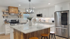 A modern kitchen with white cabinets, a stainless steel refrigerator, a central island with stools, and a pendant light. - Incredible retreat on the Guadalupe River (Hunt)