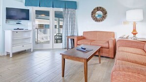 Sala de estar com mesa de madeira, dois sofás laranja, televisão em prateleira e varanda com vista.