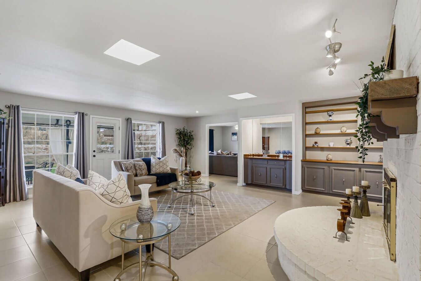 A spacious living room with a sofa, coffee table, and a glass-top table. There is a kitchen area with cabinets and a countertop visible in the background.