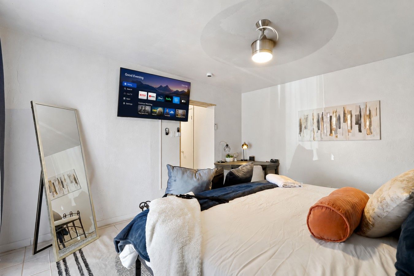 Ein modernes Schlafzimmer mit einem Bett, einem Schreibtisch, einem Stuhl, einem großen Spiegel und einem am Wand montierten Fernseher, der eine Streaming-Dienstanwendung zeigt.