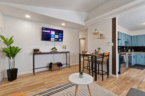 Un salon moderne comprenant un coin repas, une cuisine avec des meubles bleus et un téléviseur à écran plat fixé au mur, qui diffuse des services de streaming.