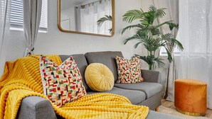 Un salon avec un canapé gris, un plaid jaune, des coussins à motifs, un pouf rond et un grand miroir au mur.