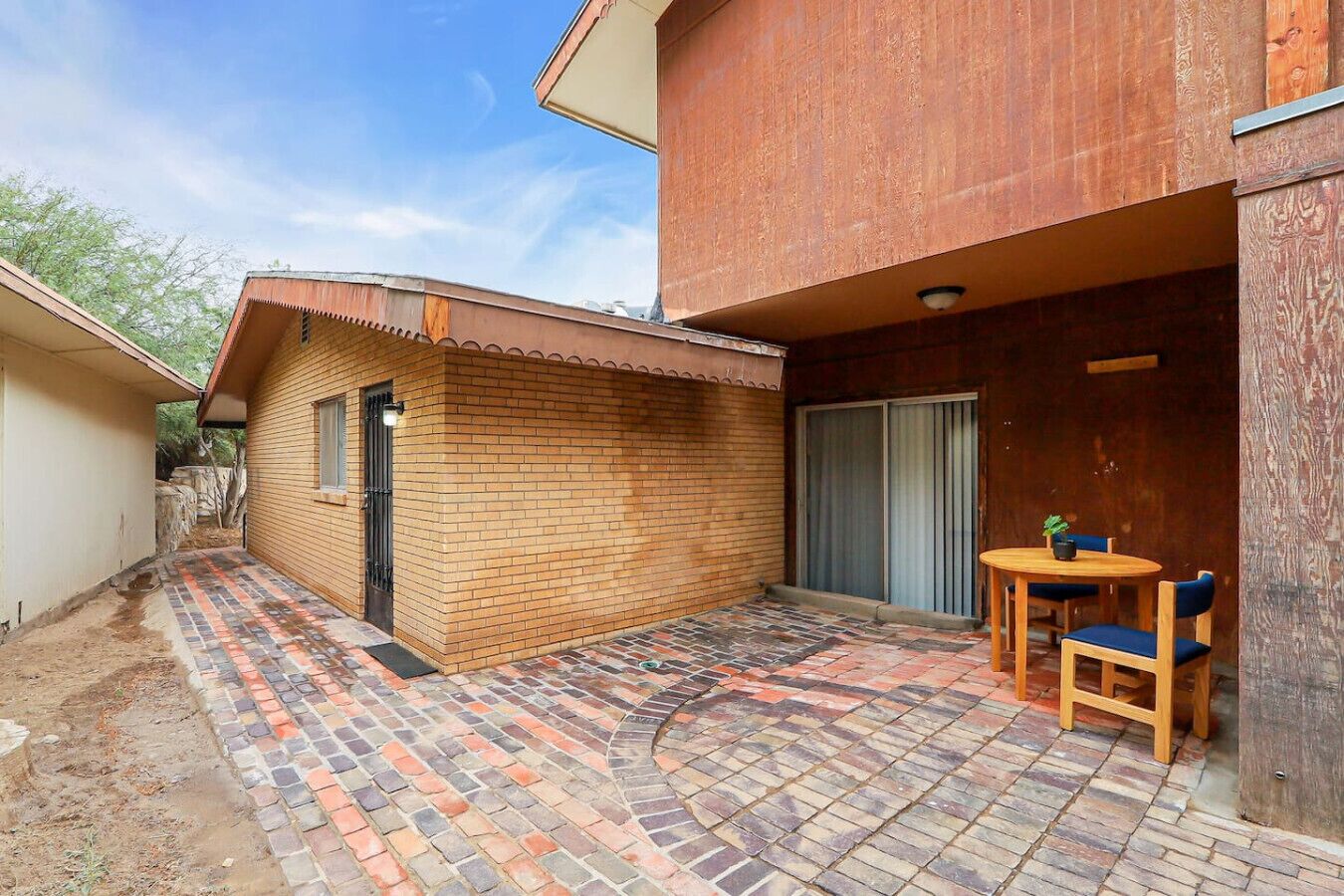 Une cour avec une terrasse en briques, un petit bâtiment en briques et une table en bois entourée de chaises.