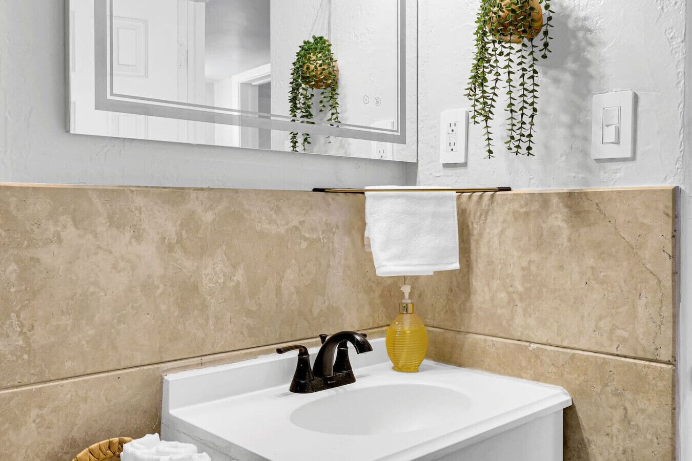 A bathroom with a white sink, black faucet, beige tiles, a mirror, and a hanging plant.