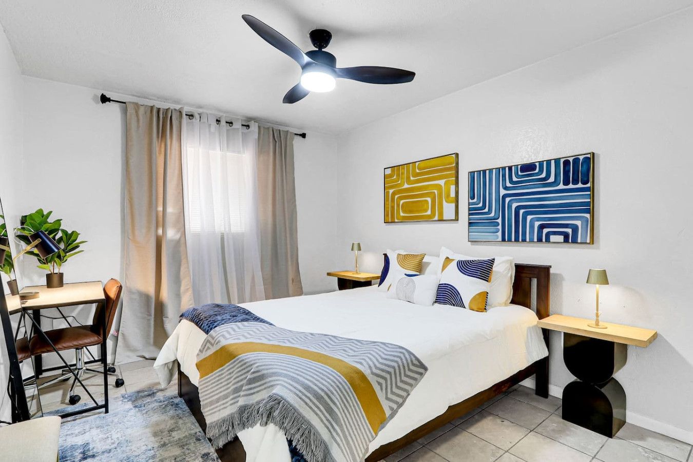 Ein modernes Hotelzimmer mit Bett, Nachttischen, einem Schreibtisch mit Stuhl, einem Deckenventilator und abstrakter Wandkunst.