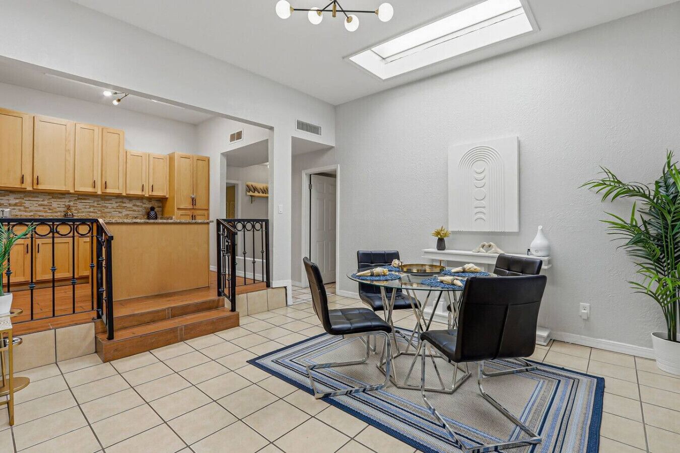 A modern kitchen with wooden cabinets, a dining area with a glass table and black chairs, and a staircase leading to an upper level.