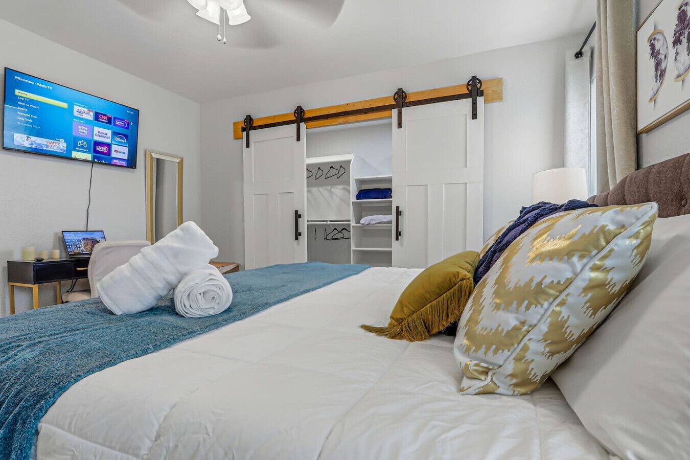 A hotel room with a bed, a TV mounted on the wall, a desk with a laptop, and a wardrobe with sliding doors.