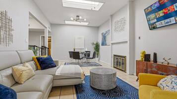 Un salon moderne avec un canapé, un pouf et un fauteuil jaune. On y trouve une table à manger avec des chaises, et un téléviseur fixé au mur.