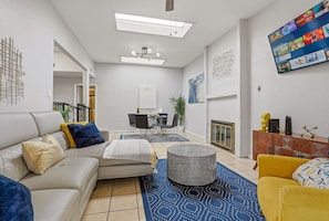 Una sala moderna con sofá, reposapiés y sillón amarillo. Hay una mesa de comedor con sillas y un televisor montado en la pared.