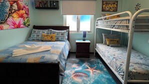 Une chambre avec un lit superposé, un lit simple, un ventilateur de plafond, une fenêtre avec des stores et une fresque murale.