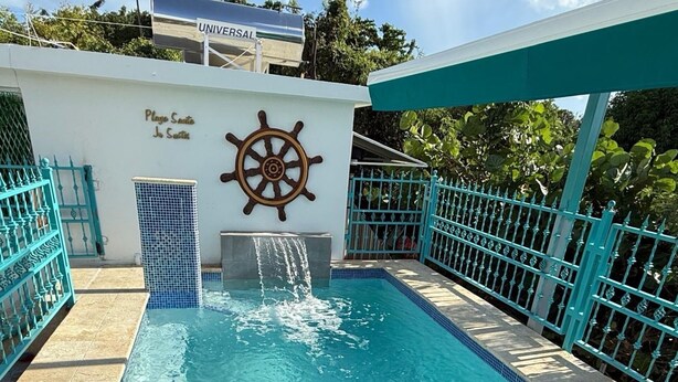 Une piscine décorée avec un gouvernail de bateau, une pancarte indiquant « Play Santos », et une enseigne « UNIVERSAL » en arrière-plan.