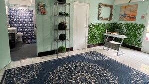 Une pièce avec un tapis à motifs, une étagère en métal ornée de plantes, une porte blanche et un mur vert agrémenté d’une bordure de plantes.