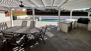 Une terrasse extérieure couverte avec une piscine, un espace barbecue et une table à manger avec des chaises.