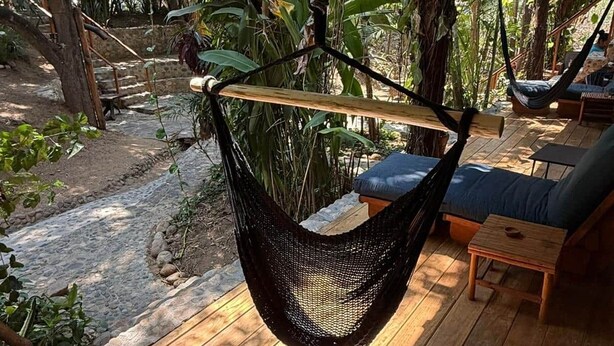 Un fauteuil suspendu en forme de hamac, accroché à une structure en bois, une table en bois et un banc rembourré offrant une vue sur un jardin et des arbres.