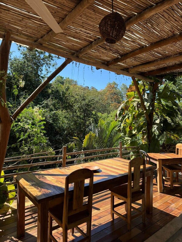 An outdoor wooden deck with tables and chairs, surrounded by lush greenery and a thatched roof structure.