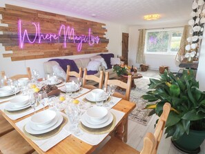 A dining area with a wooden table set for a meal, a neon sign reading "Where Magic Lives", and a cozy living space in the background. - Piskies Magic Home (Penryn)