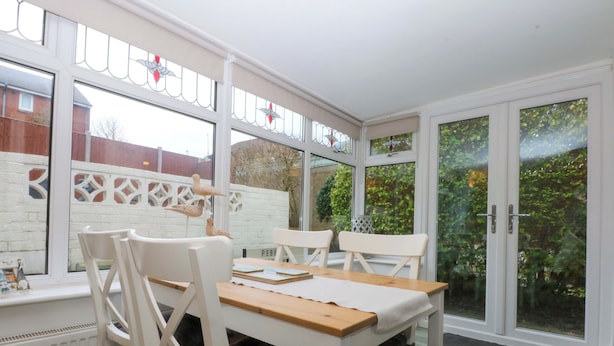 Uma área de refeições com cadeiras brancas e uma mesa de madeira, janelas amplas com vidro decorativo e vista para o verde lá fora.