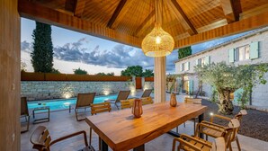 An outdoor patio with a pool, wooden furniture, and a hanging light fixture.