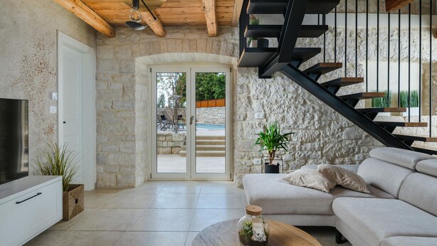 A modern living room with a stone wall, a wooden ceiling, a staircase, and a view of an outdoor swimming pool.