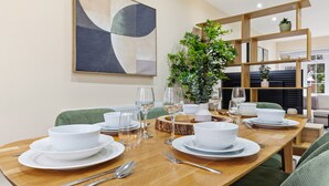 Comedor con una mesa de madera dispuesta para cuatro, con platos, cuencos y cubiertos blancos. Incluye sillas tapizadas de verde, una planta en maceta y un estante de madera con objetos decorativos.