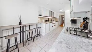 Een moderne keuken met witte kasten, een marmeren aanrechtblad en een gasfornuis. Er staan drie barkrukken en een radiator onder het aanrecht.