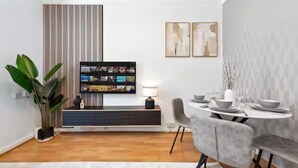 Een moderne eetruimte met een ronde tafel, grijze beklede stoelen, een zwarte entertainmentkast en een potplant.