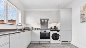 A modern kitchen with white cabinets, a built-in oven, a washing machine, and a window with blinds. - Fast WiFi & 5 Beds | Group Getaway | Sleeps 7 (Greater London)