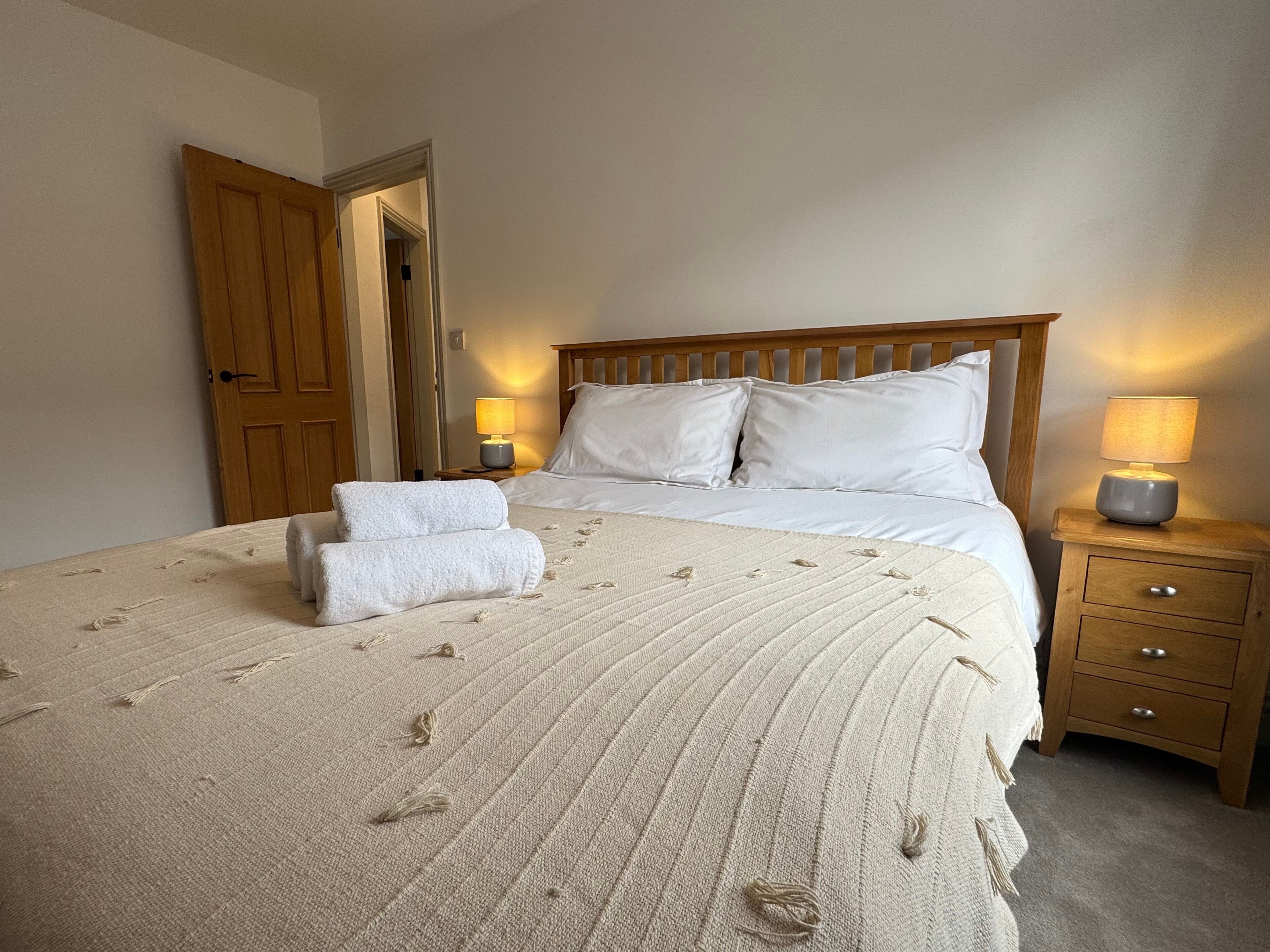 A neatly made bed with a quilt, two pillows, and a pair of towels. There are two bedside lamps and a wooden nightstand with drawers.