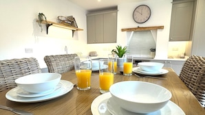 Une table à manger est dressée avec des bols, des assiettes et des verres remplis de jus d’orange, le tout dans une cuisine moderne dotée de meubles en osier et d’une horloge au mur.