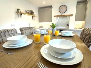 A dining table set with white bowls, plates, and glasses of orange juice in a modern kitchen with wicker furniture and a clock on the wall. - Vine Cottage - 3 Bedroom, Stamford Centre. (Lincolnshire)