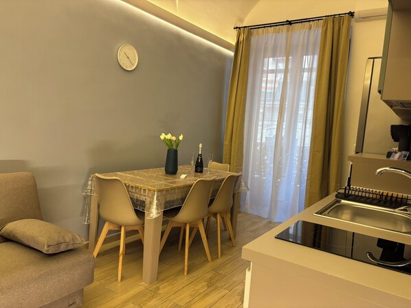 A compact living space with a dining table, chairs, a sofa, a clock, a window with curtains, and a kitchen area with a sink and a coffee machine.