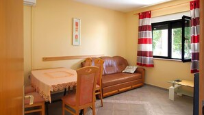 Un espacio habitable compacto con sofá, una mesita y una silla. Se ve una ventana con cortinas, y un cuadro cuelga en la pared.
