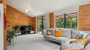 A modern living room with a wooden interior, a sofa set, a TV, and a large window offering a view of greenery. - Whare Marie - Direct Redwoods Trail Access! (Rotorua)