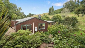 A wooden cabin with a satellite dish, surrounded by lush greenery and ferns. - Whare Marie - Direct Redwoods Trail Access! (Rotorua)