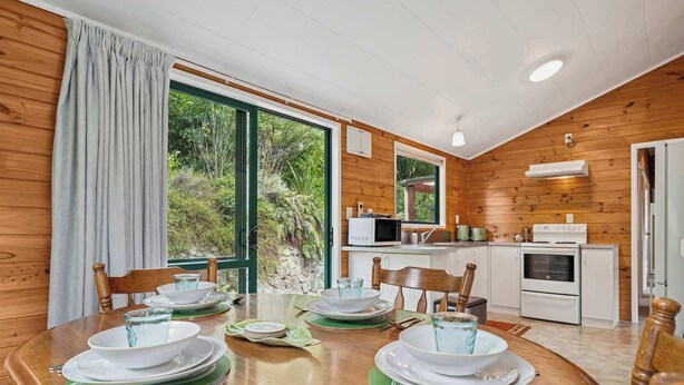 A dining area with a wooden table set for four, a kitchen with modern appliances, and large windows offering a view of greenery. - Whare Marie - Direct Redwoods Trail Access! (Rotorua)