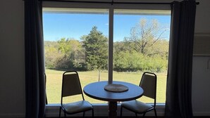 A room with a round table and two chairs, a window with curtains, and a view of a grassy area with trees.