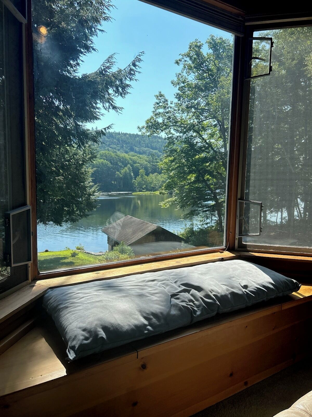 Une pièce dotée d’une grande fenêtre donnant sur un lac, d’un banc en bois avec un coussin, et offrant une vue sur une zone boisée.