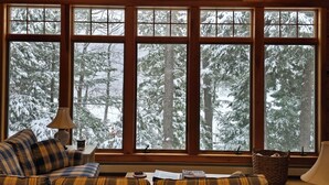 A cozy cabin interior with a large window overlooking a snowy forest. There is a plaid sofa, a lamp, and a basket on the windowsill. - Stunning Belgrade Lakes Waterfront Retreat – Dock, Kayaks & Breathtaking Views! (Rome)
